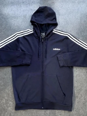 Svart Adidas hoodie med dragkedja - Klassisk svart hoodie från Adidas med vit logga på bröstet och tre vita ränder längs ärmarna. Hoodien har dragkedja framtill, justerbar huva med snören och två stora fickor. Perfekt för en sportig och avslappnad stil.