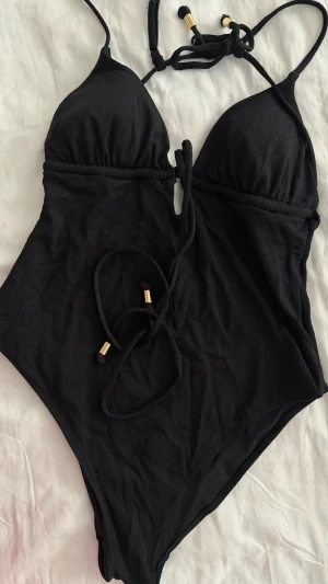 Svart baddräkt med knyt från H&M - Stilren svart baddräkt från H&M med knyt i midjan och smala axelband med guldfärgade detaljer. Baddräkten har vadderade kupor. Perfekt för stranden eller poolen. Oanvänd säljs för halva priset 