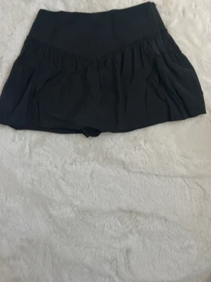 Svart volangkjol i lätt material - Svart kortkjol med volangdetaljer och bred midja. Kjolen är gjord i ett lätt och luftigt material som ger en skön känsla. Perfekt att styla med en crop top eller oversized hoodie för en trendig look. Det finns även innebyxor/shorts för att du ska känna dig säker och trygg