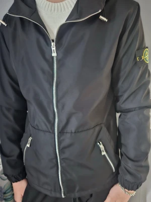 Svart vindjacka från Stone Island - Snygg svart vindjacka från Stone Island med dragkedja framtill, två snedställda fickor med dragkedja och klassisk logotyp-patch på vänster ärm. Jackan har en sportig look och är tillverkad i ett lätt, vindtätt material.