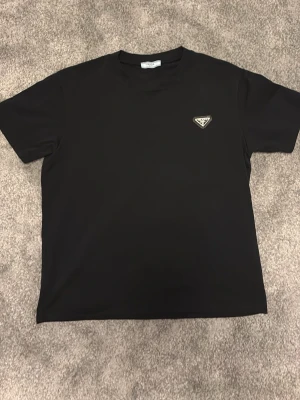 Svart t-shirt från Prada - Snygg svart t-shirt från Prada med klassisk passform och diskret logga på bröstet. T-shirten har rund halsringning och korta ärmar, perfekt för en stilren och trendig look.