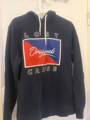 Blå hoodie från Jack & Jones - Mörkblå hoodie från Jack & Jones Originals med tryck i rött, blått och vitt framtill där det står 'Lost Cause Originals'. Huva med vita dragsnören och ribbade muddar. Perfekt för en avslappnad stil.