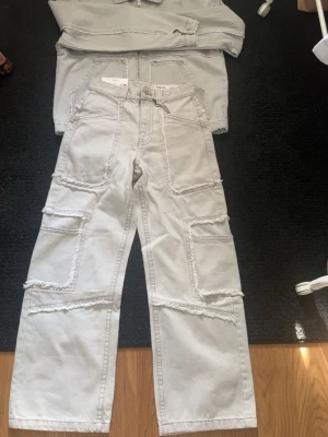 Vita cargobyxor från Bershka - Snygga vita cargobyxor från Bershka med raka ben och flera stora fickor på sidorna. Byxorna har råa kanter och detaljerade sömmar som ger en cool och trendig look. Perfekta för dig som gillar streetstyle och vill sticka ut lite extra.