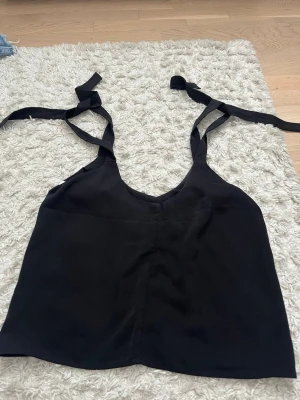 Svart linnetopp med knytband - Stilren svart linnetopp med v-ringning och breda axelband som knyts upptill. Toppen är ärmlös och har en rak passform, perfekt för en trendig och enkel look. Materialet känns mjukt och lätt mot huden.
