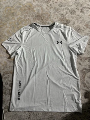 Vit Under Armour t-shirt i polyester - Snygg vit t-shirt från Under Armour med svart logga på bröstet och tryck på ärmen och sidan. Tillverkad i lätt och funktionell polyester, perfekt för träning. Klassisk rund hals och korta ärmar för en sportig look.