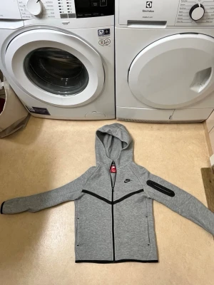 Grå Nike hoodie med dragkedja - Barn storlek Snygg grå hoodie från Nike med hel dragkedja och svarta detaljer. Huva och praktisk ficka med dragkedja på ena ärmen. Perfekt för en sportig och avslappnad stil. Nike-logga på bröstet och liten logga bak. Mjuk och bekväm bomullsblandning.