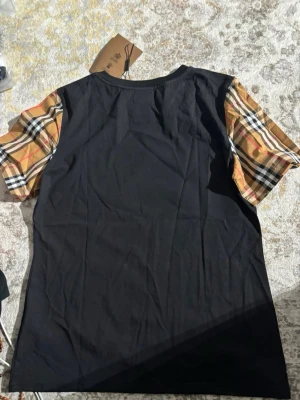 Svart Burberry t-shirt med rutiga ärmar - Snygg svart t-shirt från Burberry med klassiska beige och rödrutiga ärmar. T-shirten har rund halsringning och är tillverkad i mjuk bomull. Perfekt för dig som vill ha en stilren look med en touch av lyx.