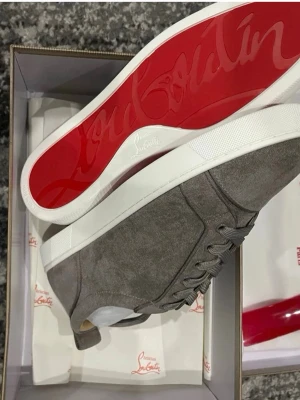 Gråa Louboutin sneakers med röd sula - Snygga sneakers från Christian Louboutin i grå mocka med klassisk röd sula och vita detaljer. Skorna har lågt skaft, tonade skosnören och stilren design. Perfekta för dig som vill sticka ut med exklusiva detaljer och lyxig känsla.