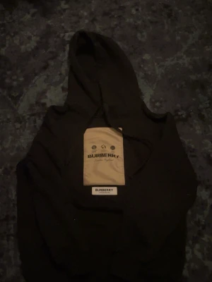 Svart hoodie från Burberry - Snygg svart hoodie från Burberry med beige patch och logga på bröstet. Klassisk huva med dragsko och ribbade muddar vid ärmslut och nederkant. Perfekt för dig som vill ha en stilren och exklusiv look.
