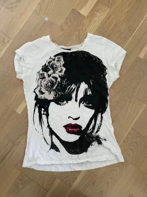 Vit t-shirt med tryck från Gina Tricot - Säljer en vit t-shirt från Gina Tricot med svart tryck av ett ansikte och rosor på framsidan. T-shirten har rund halsringning och korta ärmar. Materialet är mjuk bomull som känns skönt mot huden. Perfekt för dig som gillar coola prints och streetstyle.