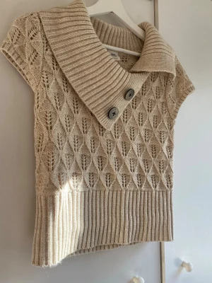 Beige stickad topp  - Beige stickad topp med korta ärmar, bred ribbad krage och två stora knappar framtill. Toppen har ett snyggt mönster i stickningen och bred ribbad nederkant. Perfekt för lager-på-lager och en trendig look.