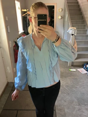 Ljusblå blus med volanger - Snygg ljusblå blus med volangdetaljer framtill och längs ärmarna. Blusen har en v-ringad hals och långa puffiga ärmar med resår vid handlederna. Perfekt att matcha med jeans eller kjol för en trendig look. Finns smala snören framtill som kan användas för att knyta en rosett eller liknande.