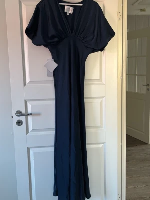Marinblå långklänning från Azazie - Elegant marinblå långklänning från Azazie Atelier med djup v-ringning och vida korta ärmar. Klänningen har en smickrande passform och är tillverkad i ett glansigt, mjukt material som ger en lyxig känsla. Dragkedja på vänsterkant. Perfekt för speciella tillfällen. 