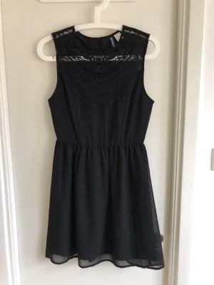 Svart spetsklänning från H&M - Svart ärmlös klänning från H&M med vacker spetsdetalj och broderier upptill. Klänningen har rund halsringning och lätt utställd kjol i kort modell. Perfekt för dig som gillar en elegant och stilren look.