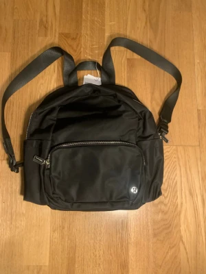 Svart ryggsäck från Lululemon - Snygg svart ryggsäck från Lululemon med justerbara axelband och en stor ytterficka med dragkedja. Tillverkad i slitstarkt nylonmaterial och har en metalllogga framtill. Perfekt storlek för vardagens alla grejer och enkel att matcha med olika outfits.