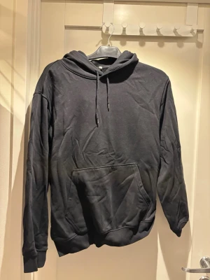 Svart hoodie från Weekday - Svart hoodie från Weekday med klassisk känguruficka och dragsko i huvan. Mjuk insida och ribbade muddar vid ärmslut och nederkant. Perfekt för en chill och avslappnad stil.