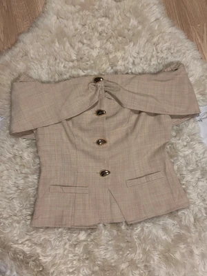 Elegant topp - Trendig beige offshoulder topp med stora guldknappar framtill och markerad midja. Snyggt omlottliknande över axlarna och två dekorativa fickor framtill. Perfekt för dig som vill sticka ut med en elegant och modern look. ‼️Oanvänd