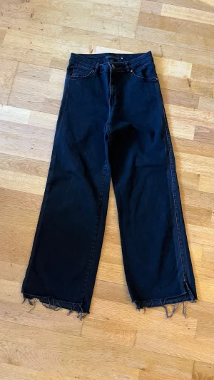 Svarta vida jeans - Säljer ett par svarta jeans med breda ben och råa, fransiga benslut. Jeansen har klassisk femficksdesign och hög midja. Perfekta för en avslappnad och trendig look. Materialet är kraftig denim i bomull.