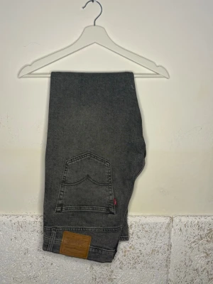 Grå Levi's jeans med rak passform - Säljer ett par grå jeans från Levi's med klassisk rak passform. Jeansen har en bakficka med den ikoniska röda Levi's-taggen och en brun läderpatch vid linningen. Tillverkade i slitstark denim, perfekta för en avslappnad och trendig look.