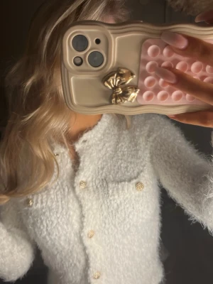 Vit fluffig kofta med guldknappar - Superfin vit kofta i fluffigt material med dekorativa guldfärgade knappar framtill. Koftan har lång ärm och en klassisk krage, perfekt för att höja vilken outfit som helst. Passar dig som gillar en chic och mysig stil.