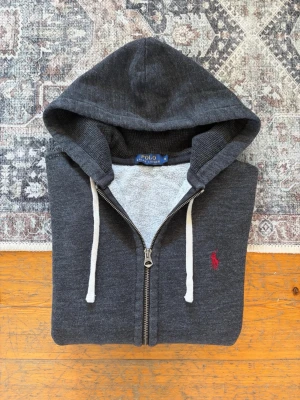 Melerad Mörkgrå Polo Ralph Lauren Zip Up Hoodie - Storlek S - Hej! 👋 Säljer en riktigt snygg Polo Ralph Lauren zip up hoodie i storlek S (Herr). Färgen är en klassisk och unik melerad mörkgrå (charcoal heather) med snygg mörkblå pony-logga på bröstet 🐎 Tröjan är i mycket gott skick med den sköna, mjuka fleece-insidan kvar ✨ Grym passform (personen på bilden är ca 177cm/67kg) 👕 Denna modell saknar QR-kod, men alla invändiga tvättlappar sitter kvar ✅ Perfekt basplagg till garderoben. Fraktar snabbt! 🚛📦💬💯 