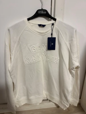 Vit sweatshirt från GANT - Snygg vit sweatshirt från GANT med broderad text 'GANT MCMXLIX' på bröstet. Tröjan har rund halsringning, ribbade muddar och är tillverkad i mjukt material som känns skönt mot huden. Perfekt för en avslappnad och stilren look. Aldrig använd prislapp fortfarande kvar. Står storlek XXL men kan tänka mig att den är liten i storlek så kanske XL