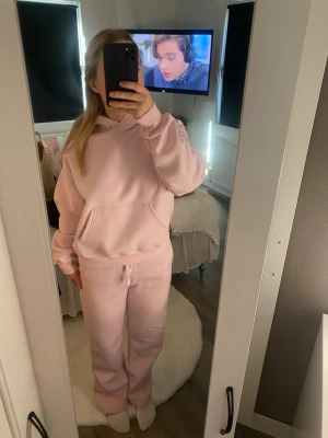 Ljusrosa mjukisset med hoodie - Mysigt loungewear-set i ljusrosa med matchande hoodie och byxor. Hoodien har stor ficka framtill byxorna har resår i midjan. 