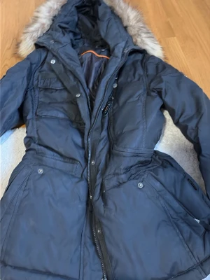 Svart parkas från Hollies med fuskpäls - Snygg svart parkas från Hollies med avtagbar huva och fuskpäls. Jackan har dragkedja, knappar, flera fickor och en markerad midja. Perfekt för kalla dagar och riktigt varm. Klassisk modell som funkar till det mesta.