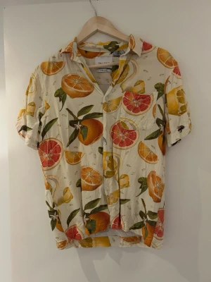 Mönstrad skjorta med citrusfrukter - Kortärmad skjorta från Vailent med färgglatt citrusmönster i orange, gul och röd på en ljus botten. Skjortan har klassisk krage, knappar framtill och en avslappnad passform. Perfekt för dig som gillar statement-plagg och somriga vibes.