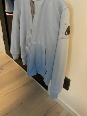 Ljusblå hoodie från Moose Knuckles - Snygg ljusblå hoodie från Moose Knuckles med dragkedja, fickor och en ikonisk logga på ärmen. Insidan är fodrad med mjuk päls och huvan är stor och skön. Perfekt för dig som vill ha en stilren och bekväm hoodie med exklusiv känsla. Funkar även som jacka 