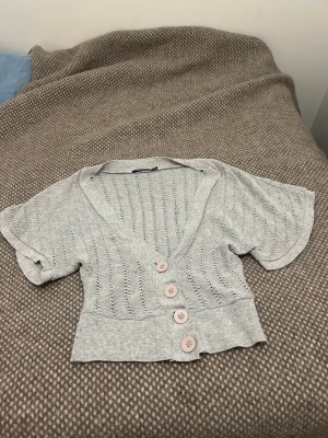 Ljusgrå kortärmad kofta Atmosphere - Säljer en ljusgrå, stickad kofta från Atmosphere med korta ärmar och djup V-ringning. Koftan har fyra stora ljusrosa knappar framtill och ett luftigt, hålmönstrat tyg. Perfekt för lager på lager och en chill look. Köp secondhand i Spanien.