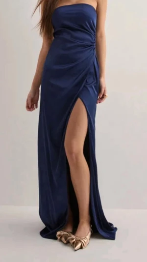 Mörkblå axelbandslös maxiklänning - Min väldigt fina och populära balklänning som är en mörkblå axelbandslös med draperad detalj i sidan och hög slits framtill. Klänningen har en elegant, figurnära passform och är tillverkad i ett glansigt, mjukt tyg som ger en lyxig känsla. 