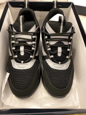 Svarta D**r b22 med grå detaljer - Snygga svarta sneakers med grå och vita detaljer. Ovandelen är i mesh och syntetmaterial för bra andningsförmåga och komfort. Skorna har en sportig look med reflekterande detaljer och klassisk snörning. Perfekta för dig som gillar stilrena och moderna sneakers.