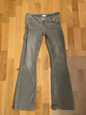 Grå bootcut jeans med fickdetalj - Säljer ett par grå bootcut jeans med låg midja och snygga fickdetaljer bak, inklusive lock med knapp. Jeansen har klassisk femficksdesign och är tillverkade i mjukt denimtyg. Perfekta för en avslappnad och trendig look.
