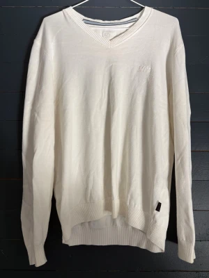 Vit stickad tröja från Hugo Boss - Klassisk vit stickad tröja från Hugo Boss med V-ringning och diskret broderad logga på bröstet. Tröjan har ribbade muddar vid ärmslut och nederkant samt en liten svart etikett i nederkant. Perfekt för en clean och stilren look.