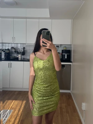 Grön glittrig kort klänning - Säljer en snygg grön glittrig kort klänning med smala axelband och figurnära passform. Klänningen har en öppen rygg och är perfekt för dig som vill sticka ut. Materialet är glittrigt och stretchigt för en bekväm känsla.