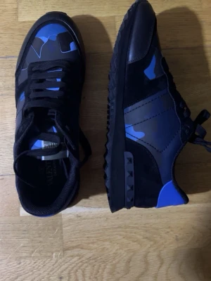 Valentino blå camo sneakers - Snygga sneakers från Valentino med blå och svarta camo-detaljer. Skorna har svarta mocka- och läderpaneler, samt en unik nubbad sula och blå häl. Klassisk låg modell med svarta skosnören och cool design som sticker ut.