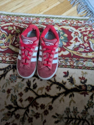 Röda Adidas sneakers med mocka - Snygga röda Adidas sneakers i mocka med klassiska vita ränder på sidorna och vita sulor. Skorna har röda skosnören och låg profil. Perfekta för dig som gillar retrostil och streetwear.