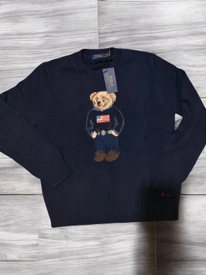 Mörkblå stickad tröja Polo Bear - Mörkblå stickad tröja från Polo Ralph Lauren med den ikoniska Polo Bear på bröstet. Björnen har en tröja med amerikanska flaggan och jeans. Tröjan har rund halsringning och ribbade muddar. Perfekt för dig som gillar klassisk preppy stil.