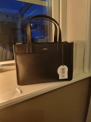 Svart handväska från DonDonna - Stilren svart handväska från DonDonna i slätt konstläder. Väskan har två handtag och medföljande axelrem med guldfärgade detaljer. Den har två innerfack, en med dragkedja och en utan. Aldrig använd. Nypris 500. Kom gärna med prisförslag