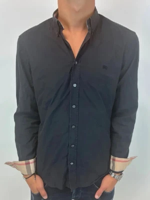 Svart skjorta från Burberry - Klassisk svart skjorta från Burberry med långa ärmar och knappar framtill. Ikoniskt rutigt tyg syns på insidan av manschetterna. Skjortan har en normal passform och diskret logga på bröstet. Perfekt för en stilren look.