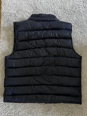 Svart dunväst med hög krage - Stilren svart dunväst med quiltad design och hög krage. Västen har breda horisontella kanaler och är tillverkad i ett glansigt material som ger en modern look. Perfekt att slänga över en hoodie eller tröja för extra värme.