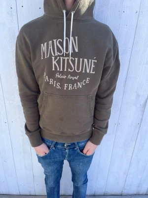 Maison Kitsuné  - Snygg brun hoodie från Maison Kitsuné med vit text på bröstet där det står 'Maison Kitsuné Palais Royal Paris, France'. Klassisk känguruficka framtill, vita dragsnören och huva. Perfekt för en chill och avslappnad stil.