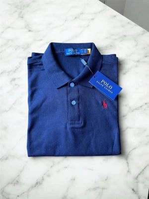 Marinblå pikétröja från Ralph Lauren - Klassisk marinblå pikétröja från Ralph Lauren med krage, knappar framtill och den ikoniska rosa broderade loggan på bröstet. Tillverkad i bomull för en bekväm och stilren look. Perfekt till jeans eller shorts för en clean outfit.