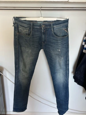 Blå slim fit jeans från Replay - Säljer ett par blå slim fit jeans från Replay med klassisk femficksmodell och snygga slitningar framtill. Jeansen har en något tvättad look och är tillverkade i bomull med stretch för extra komfort. Perfekta för en avslappnad och trendig stil.