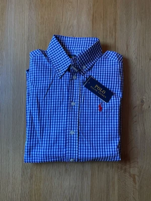Blårutig skjorta från Polo Ralph Lauren - Klassisk blårutig skjorta från Polo Ralph Lauren med vit knappstängning och röd broderad logga på bröstet. Skjortan har en snygg krage och är tillverkad i bomull för en bekväm känsla. Perfekt för dig som gillar stilrena och tidlösa plagg.