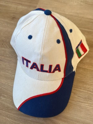 Vit och blå keps med ITALIA-brodyr - Snygg vit keps med blå och röda detaljer samt broderad texten 'ITALIA' framtill och baktill. Kepsen har även en italiensk flagga på sidan och justerbar rem bak. Tillverkad i bomullstyg och har klassisk böjd skärm.