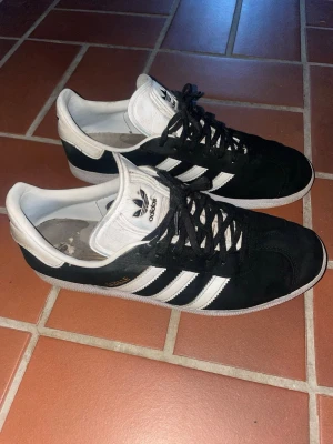 Adidas Gazelle svarta sneakers mocka - Klassiska Adidas Gazelle sneakers i svart mocka med vita detaljer och de ikoniska tre ränderna på sidan. Skorna har vit plös och häl, samt svart snörning.