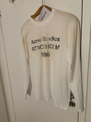 Vit långärmad topp Acne Studios - Vit långärmad topp från Acne Studios med tryckt text 'Acne Studios STOCKHOLM 196' på bröstet. Klassisk rund halsringning och relaxed fit. Tillverkad i mjuk bomull som känns skön mot huden. Perfekt för en clean och stilren look.