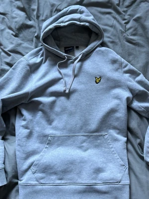 Ljusgrå hoodie från Lyle & Scott - Snygg ljusgrå hoodie från Lyle & Scott med klassisk gul logga på bröstet. Hoodien har huva med dragsko och en stor magficka framtill. Tillverkad i mjukt bomullsmaterial som är skönt att ha på sig. Perfekt för en avslappnad och stilren look. Liten o storleken
